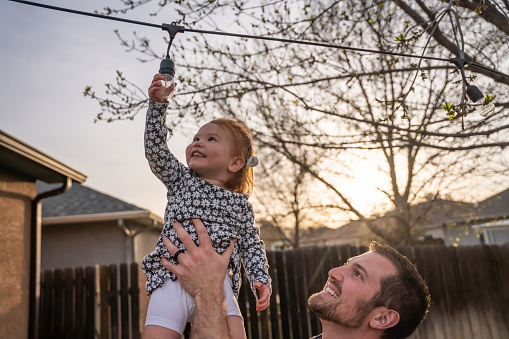 Young Father Holds Up Two-year Old daughter as she touches an edison light bulb decoratively arranged in a back yard at dusk
