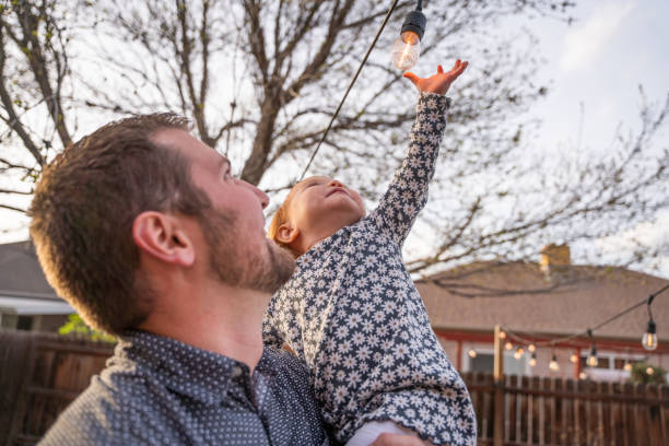 istock Young Father Holds Up Two-year Old daughter as she touches an edison light bulb decoratively arranged in a back yard at dusk 1391295355