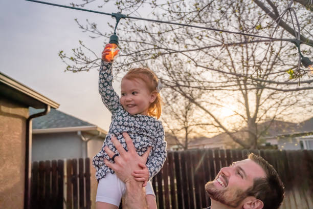 istock Young Father Holds Up Two-year Old daughter as she touches an edison light bulb decoratively arranged in a back yard at dusk 1391295372