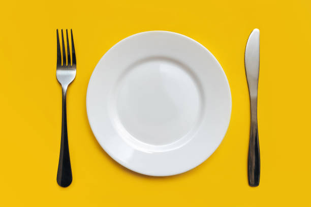Plastic excrement of a cat lies on a white plate between a fork and a knife close-up on yellow tableclothes. The concept of an inept cook, a crappy restaurant, bad food, a taste of shit. Bad smelling joke. stock photo