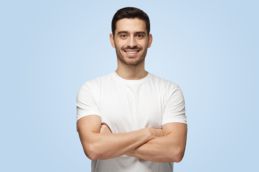 Portrait of smiling handsome man in white t-shirt standing with crossed arms isolated on blue background