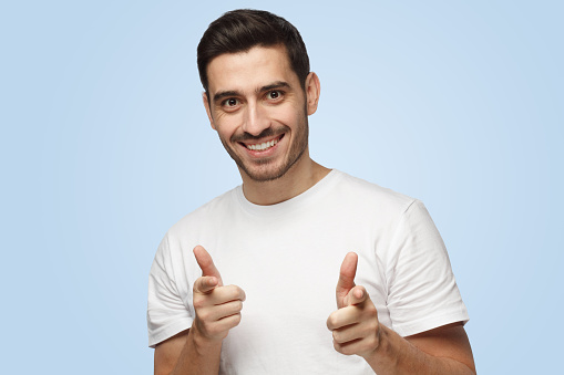 Closeup picture of good-looking European man isolated on blue background wearing white T-shirt smiling happily with open mouth and pointing to camera with both hands as if saying it is their turn