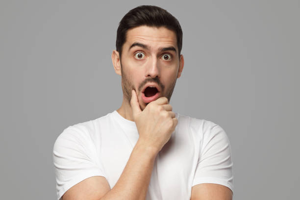 Young man with eyes wide open with fear or amazement, covering open mouth with palm Horizontal portrait of young European man isolated on grey background pictured with eyes wide open with fear or amazement, covering open mouth with palm, astounded with news wtf stock pictures, royalty-free photos & images