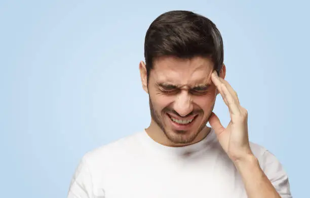 Horizontal photo of good-looking Caucasian man pictured isolated on blue background in right side of picture showing how much his head hurts, experiencing pain, looking miserable and exhausted