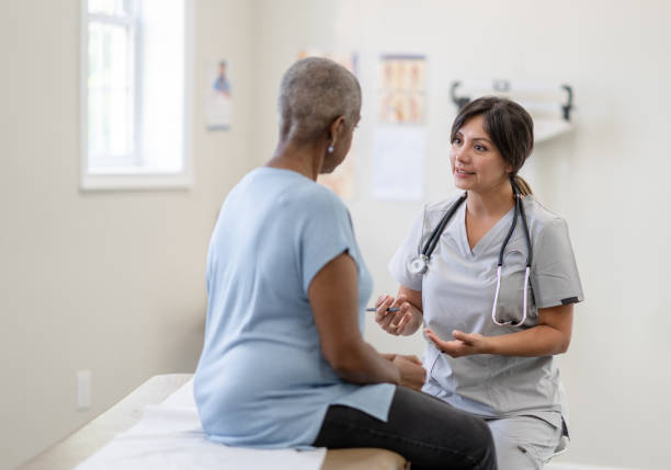 Cancer Patient Having a Check-Up A middle aged woman of African decent, who is fighting Cancer, sits up on an exam table for a check-up.  She is dressed casually and has her back to the camera.  The female nurse is seated in front of her in light grey scrubs as the two talk about how she is feeling and how to combat her side effects. woman talking to doctor stock pictures, royalty-free photos & images