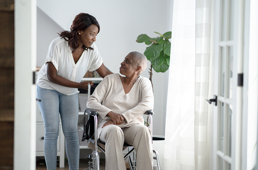 A young woman pushes her mother in a wheelchair through the house as she comes to help and support through her Cancer journey. They are both dressed in neutral tones as they look at one another.