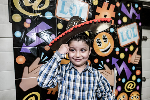 Young Mexican boy with party sombrero at the birthday party. He is wearing casual clothes with plaid shirt and jeans. Interior of car and motorcycle repair shop at night.