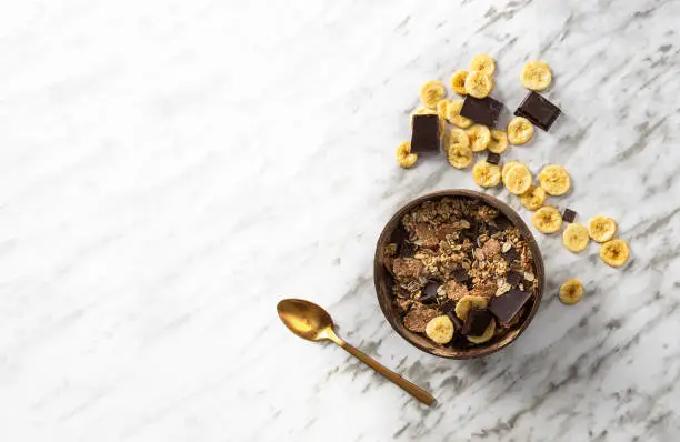 Photo of Natural muesli  in coconut bowl with  banana and chocolate on the marble table. Mix of unprocessed whole grains, chia, quinoa, nuts, seeds, fruit for heathy breakfast.