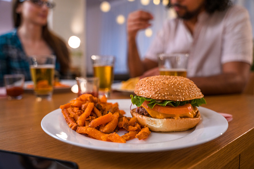 Close up on a tasty fresh looking burger with sweet potato french fries served on a plate. Diverse group of friends having dinner party.