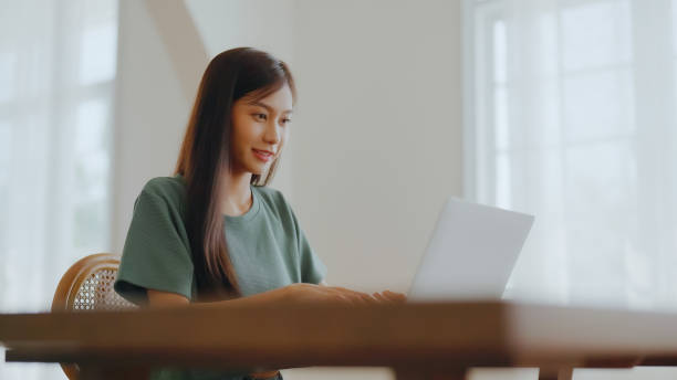 istock Smiling asian young woman working on laptop at home office. Young asian student using computer remote studying, virtual training, e-learning, watching online education webinar at house 1391337371