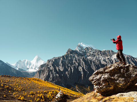 asian man standing on top of rock taking photo of Yangmaiyong (or Jampayang in Tibetan) mountain peak in the distance in Yading, Daocheng County, Sichuan Province, China