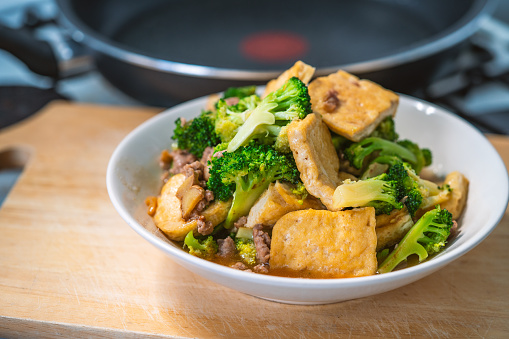 Stir-fried Broccoli and fried soft tofu, healthy Asian homemade menu, food in white plate that place on wooden cutting board in a kitchen, close up the food, natural lighting image.