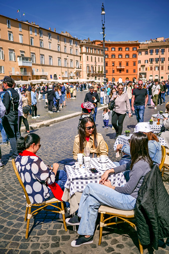 Rome, Italy, April 12 -- Some young tourists enjoy life in a sidewalk cafe and restaurant in Piazza Navona, in the historic and baroque heart of Rome. The Rione Parione, around Piazza Navona, is one of the most beautiful and visited districts of Rome for the presence of countless artistic and historical treasures, monuments and ancient Romanesque and Baroque churches, but also for its squares and alleys to be explored freely, where it is easy to find typical restaurants, pubs, small shops of artisans and scenes of daily life with the original Roman soul. In 1980 the historic center of Rome was declared a World Heritage Site by Unesco. Image in high definition format.