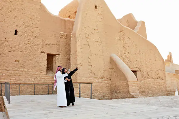 Full length view of mid adult man and woman in traditional Saudi attire standing out front Salwa Palace and capturing memories at Diriyah ruins. Property release attached.