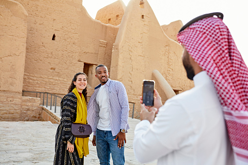 Over the shoulder view with focus on smiling mid adult couple posing for a captured memory during visit to At-Turaif open air museum near Riyadh. Property release attached.