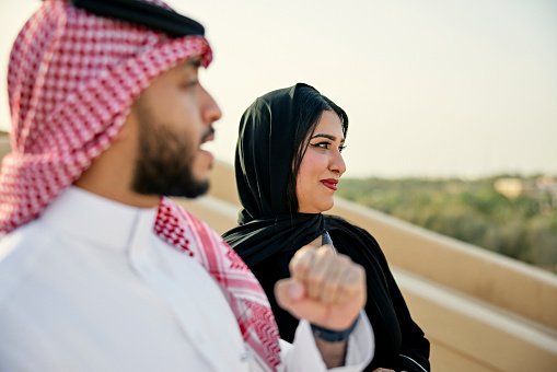 Three-quarter front view of man and woman in their 30s wearing traditional Saudi attire and looking at view of desert oasis and heritage site near Riyadh. Property release attached.