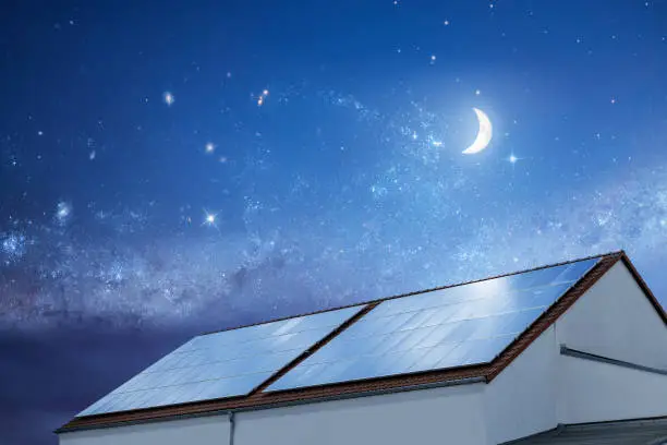 Top part of a building covered with large solar panels at night. Night sky with moon and stars in the background.