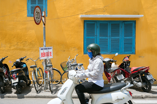Hoi An, Vietnam - May 07, 2018: Woman riding motorbike and row of parked motorbikes against yellow house.