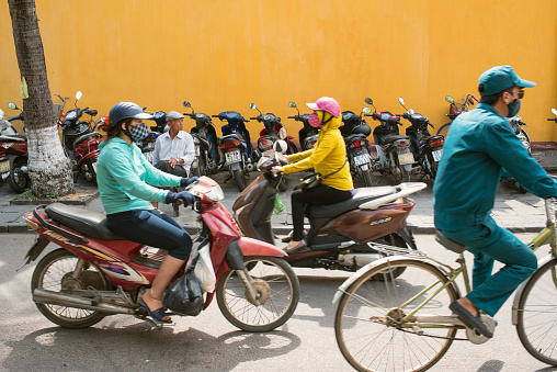 Hoi An, Vietnam - May 06, 2018: People riding motorcycle and row of parked motorcycles against yellow house wall.