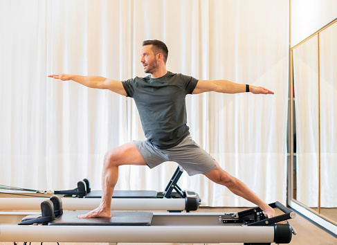 Fit man performing a lunge and stretch Warrior 2 yoga pose on a pilates reformer bed to stretch and strengthen his hip and chest muscles and promote stamina