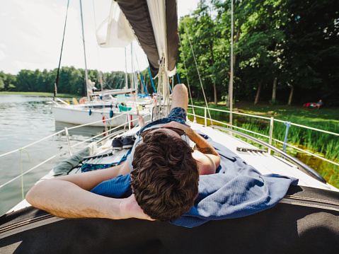 adult man laying on a sailboat deck and taking sunbath during his vacations in a sunny summer day. Yacht moored on a wooden pier on a lakeshore