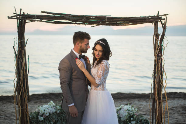 gli sposi sotto l'arco sulla spiaggia. tramonto, crepuscolo - wedding beach bride groom foto e immagini stock