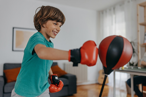 Boy is practices boxing at home, wears boxing gloves as he punches the bag very hard