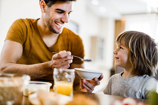 Happy father enjoying while feeding his small son with cereal during breakfast at dining table.