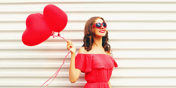 Portrait of happy smiling woman with red heart shaped balloon wearing sunglasses on white background stock photo