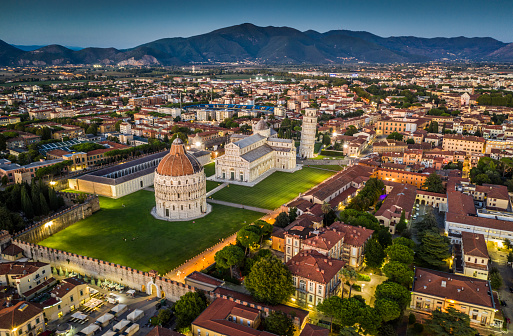 Aerial view of famous Leaning Tower of Pisa and Cathedral Santa Maria surrounded by the illuminated cityscape of Pisa at dusk. Piazza dei Miracoli, Tuscany - Italy