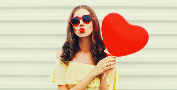 Portrait of beautiful young woman with red heart shaped balloon blowing her lips sending sweet air kiss wearing sunglasses on white background