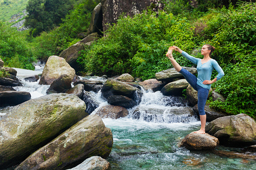 Yoga outdoors - woman doing Ashtanga Vinyasa Yoga balance asana Utthita Hasta Padangushthasana - Extended Hand-To-Big-Toe Pose position posture outdoors at waterfall