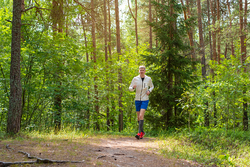 Young handsome caucasian man in a white running jacket and blue shorts running in the forest on a sunny summer day.
