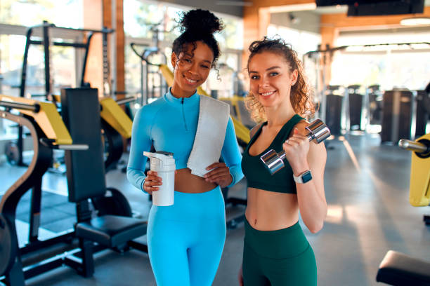 Sports and gym activities African American woman with a bottle of water in sportswear and a Caucasian woman with dumbbells standing in the gym against the background of equipment. Active healthy lifestyle, sport. community health center stock pictures, royalty-free photos & images