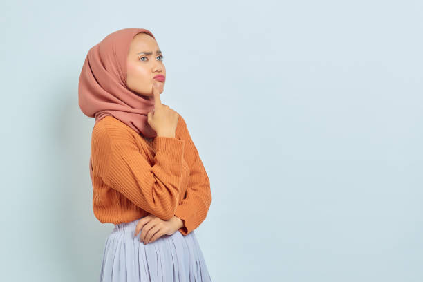 nachdenkliche junge asiatische muslimische frau in braunem pullover und hijab, die ernsthaft über eine auf weißem hintergrund isolierte frage nachdenkt - zurückhaltende kleidung stock-fotos und bilder