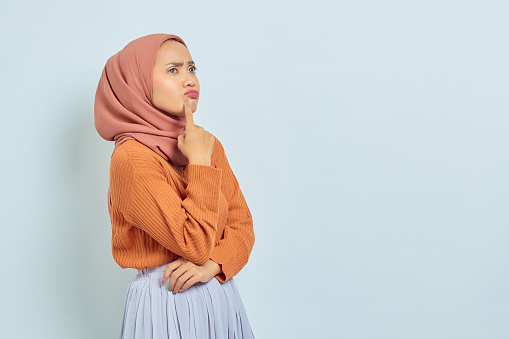 Pensativa joven musulmana asiática con suéter marrón y hiyab luciendo seria pensando en una pregunta aislada sobre fondo blanco photo