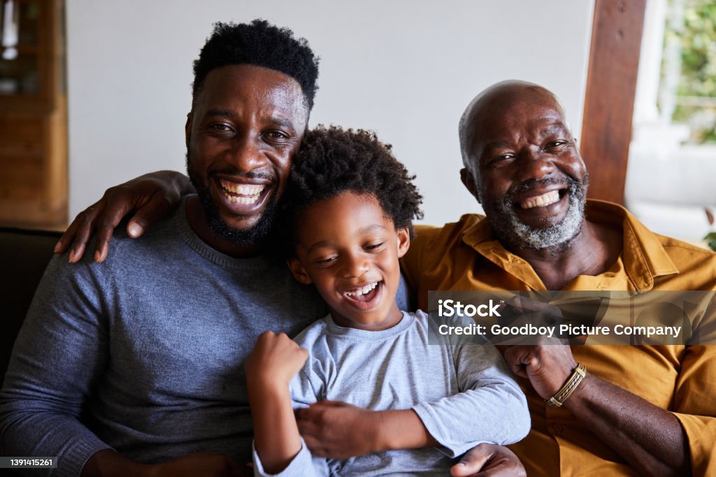 Man and his senior dad and young son laughing together on a sofa Portrait of an African man and his senior father and young son laughing while sitting on a sofa in their living room at home Father Stock Photo