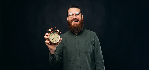 Smiling bearded man is holding an alarm clock while looking at the camera. Studio shot over black background.