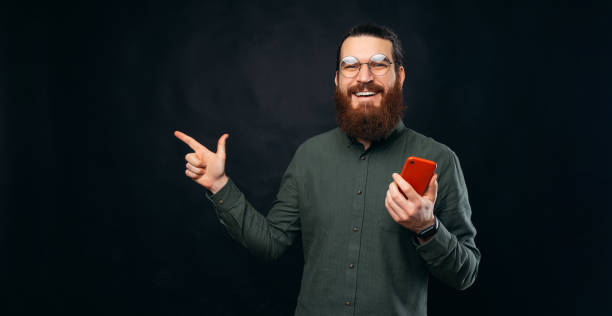 istock Smart bearded man is holding a red phone while pointing aside on backdrop. 1391418829