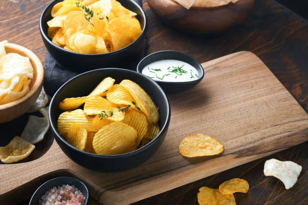 copeaux ondulés de pomme de terre. restauration rapide. bol noir en céramique croustillant de croustilles avec sauce à la crème sure et oignons dans un support en bois sur un vieux fond en bois de table de cuisine. tradition américaine. bbq chaud. vue - chips photos et images de collection