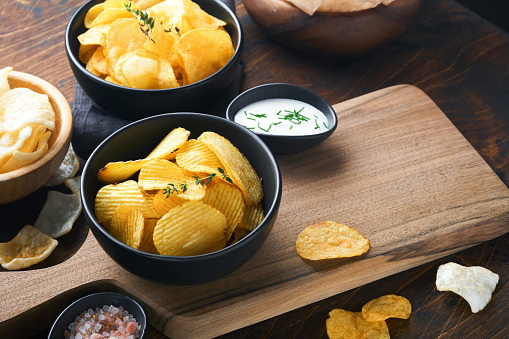 Potato corrugatedchips. Fast food. Crispy potato chips ceramic black bowl with sour cream sauce and onions in wooden stand on old kitchen table wooden background. American tradition. Hot BBQ. Top view