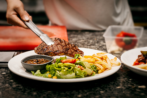 The chef puts the steak on a plate already decorated with french fries and vegetables in the kitchen