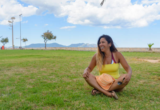 beautiful latin woman 40 years old, smiling sitting on the grass of a park in mallorca, balearic islands, hollidays concept - independence lifestyles smiling years imagens e fotografias de stock