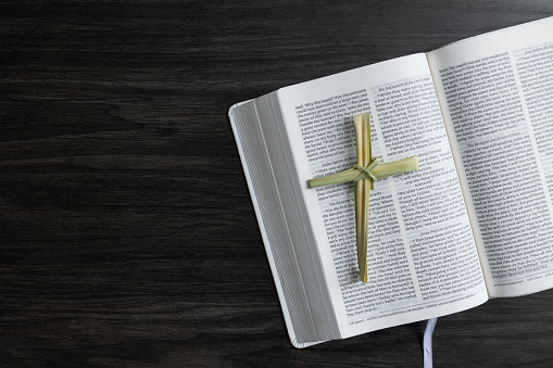 Cross made out of a dried palm leaf laying on an open bible shot from above on a dark wood table with copy space
