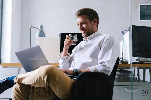 Young entrepreneur, sitting in his office with a laptop in his lap, working on it. He is holding a glass of water in one hand, while with another one is typing on the laptop.