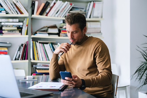 Young businessman sitting at a desk in the office. He is holding his smartphone, looking at his notebook and ideas written in it.