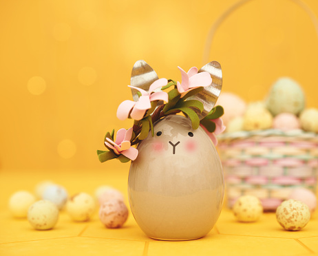 Cute gray Easter bunny decoration in a vibrant yellow setting with speckled Easter eggs and a full Easter basket