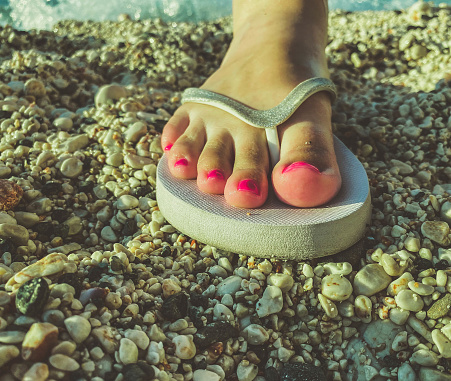 a girl with large fingers and a pink pedicure on her feet walks in a warm country in white rubber slippers. leisure shoes stands on the sand. slates for walking along the sea coast.