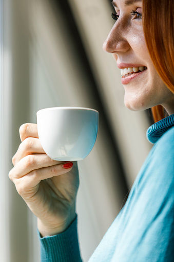 Portrait of beautiful young redhead standing by the window, enjoying a cup of coffee, looking outside, contemplating.