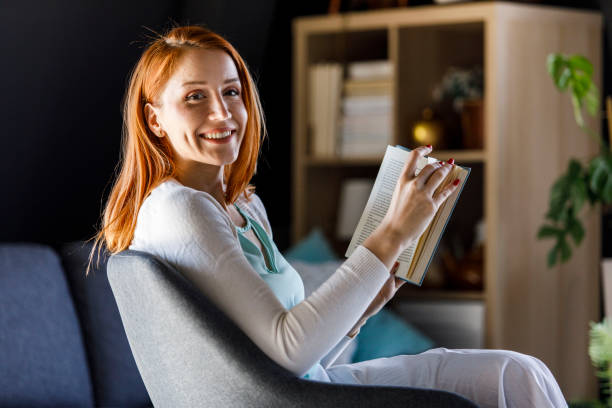 Copy space shot of joyful young redhead relaxing in her living room and enjoying reading an interesting book. She is looking at camera and smiling cheerfully.